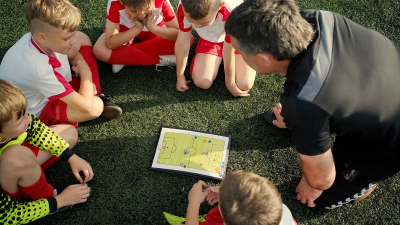 A youth soccer coach using a tactics board to explain strategy to young players seated around him