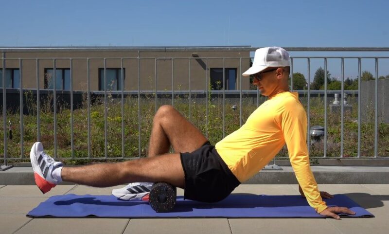 A man performs a hamstring stretch using a foam roller outdoors