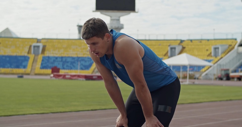 A sprinter resting at the edge of the track, visibly drained, an athlete struggling mentally