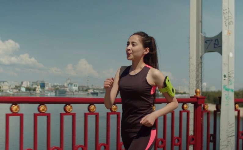 A woman walks fast along a red bridge railing, the city skyline a backdrop.
