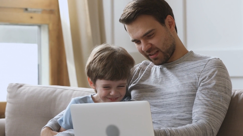 A father and son sit on a couch, smiling as they look at a laptop together