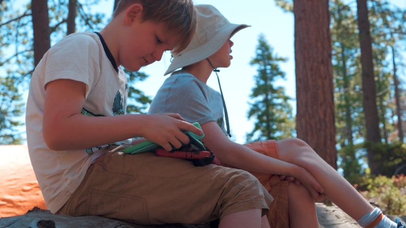 Two boys sit on a log during summer camp, one checking his gear