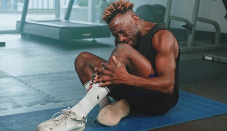 A man sits on a gym mat, clutching his injured ankle