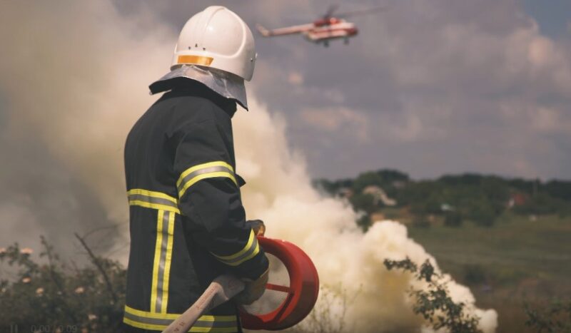 Firefighter holding a hose with a helicopter hovering in the background, ready to assist in firefighting efforts