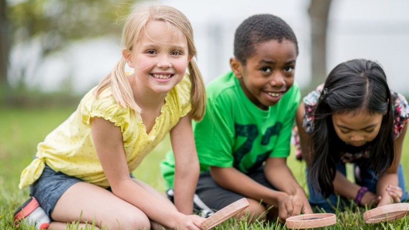 Three kids sit on the grass at summer camp, smiling and playing with wooden blocks