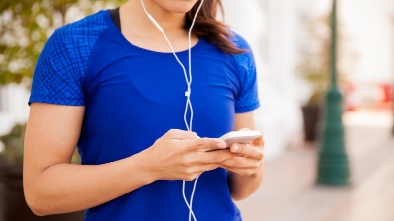 A woman in a blue shirt looks at her phone with earphones in, ready to listen to a running podcast
