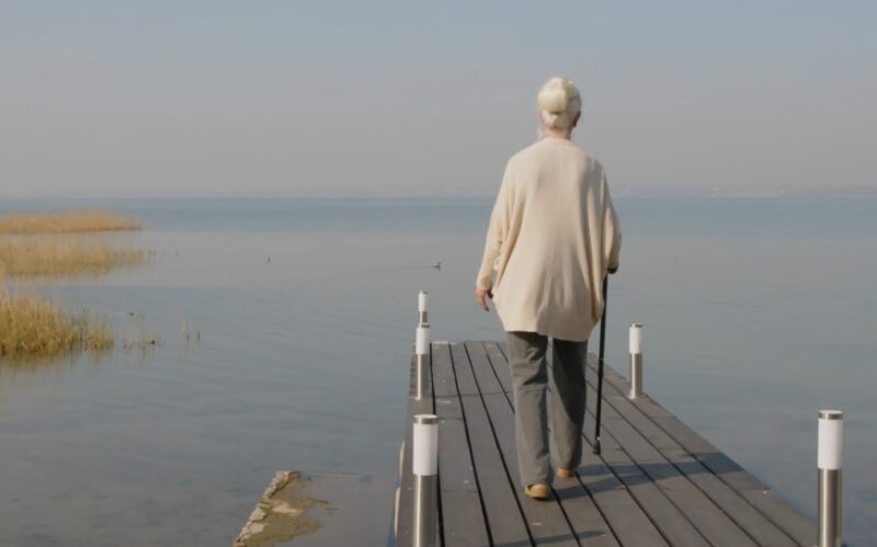 A serene elderly woman strolls along a tranquil lakeside pier
