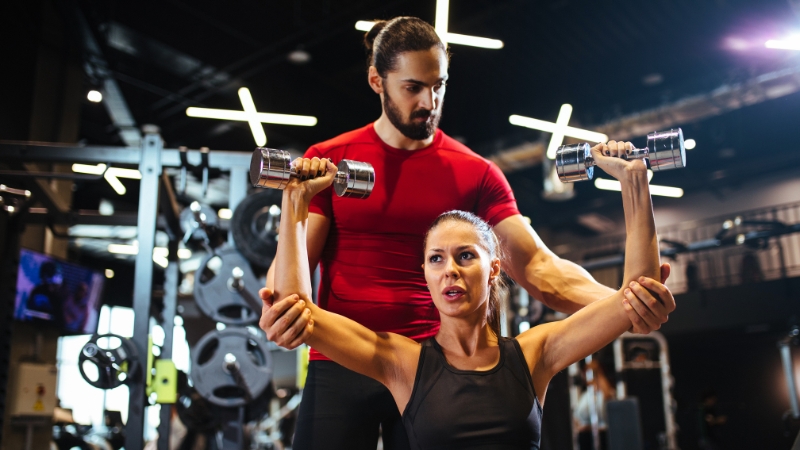 A coach supports a woman lifting dumbbells, showing calm and confidence during her workout