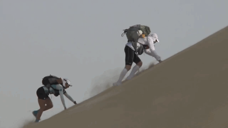 Two runners crawl up a steep sand dune under the blazing sun during an extreme desert race