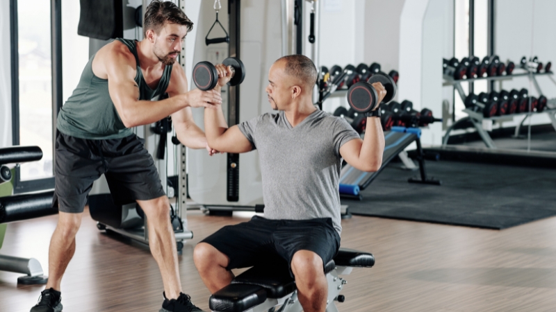 A coach guides a man through a shoulder exercise with dumbbells at the gym
