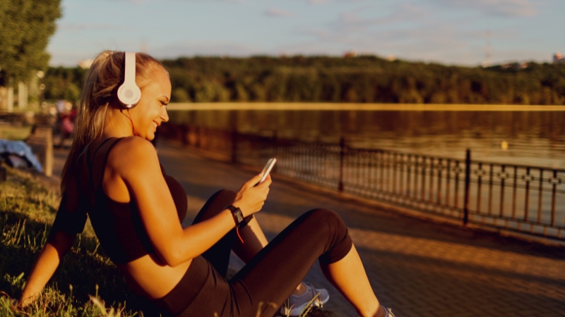 A woman sits by the water in workout clothes, smiling as she listens to a running podcast on her phone