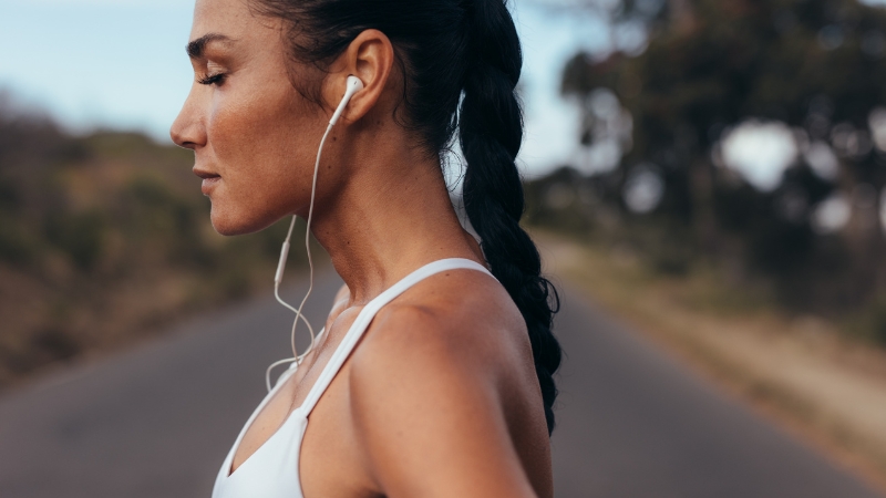 A woman with a braid listens to a running podcast through earphones while standing on a quiet road