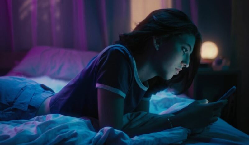 A young woman engrossed in her phone, bathed in the cool glow of a bedroom's vibrant lighting