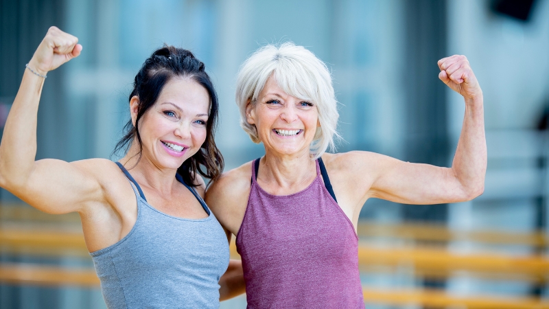 Two smiling women flex their arms, showing strength and confidence as part of a fitness after 50 lifestyle