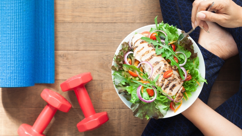 A person sits cross-legged with a fresh chicken salad, next to dumbbells and a yoga mat, showing balance between food and fitness goals