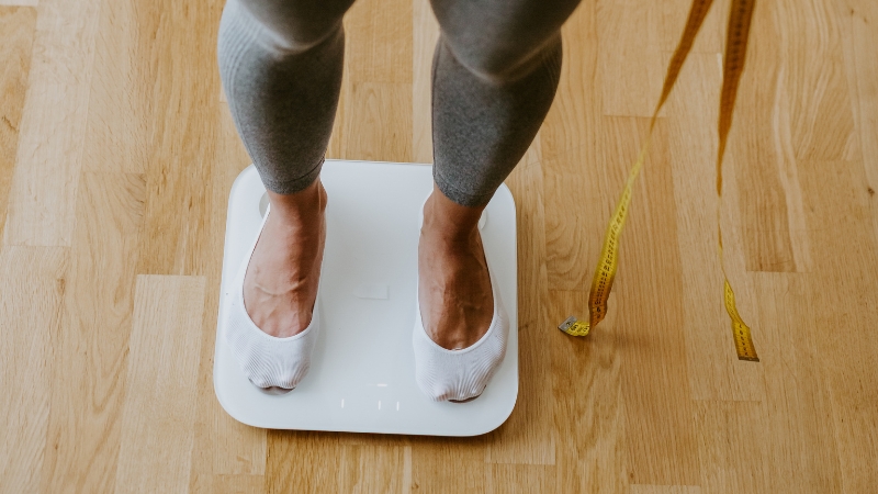 A woman stands on a digital scale with a tape measure nearby, checking her ideal weight range for her 30s