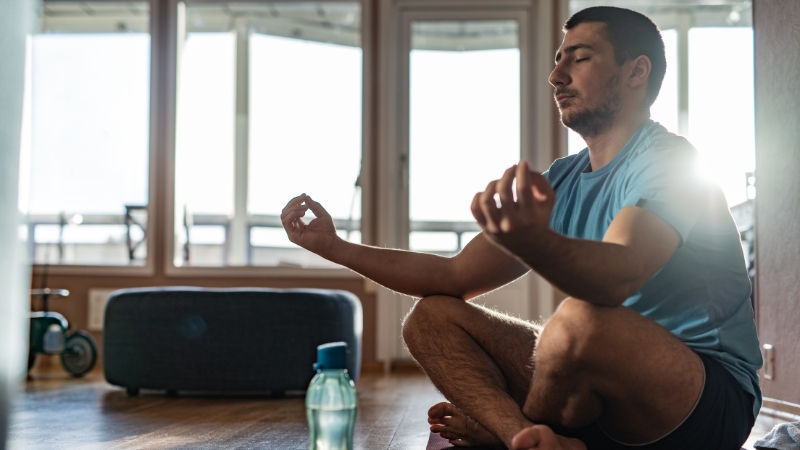 A man sits cross-legged on a yoga mat indoors, eyes closed, meditating