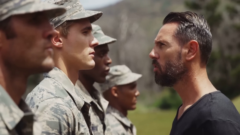 A military instructor stands face-to-face with soldiers in uniform during outdoor training