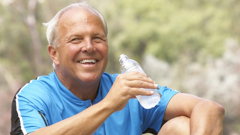A smiling older man in a blue shirt holds a water bottle during a break from exercise outdoors