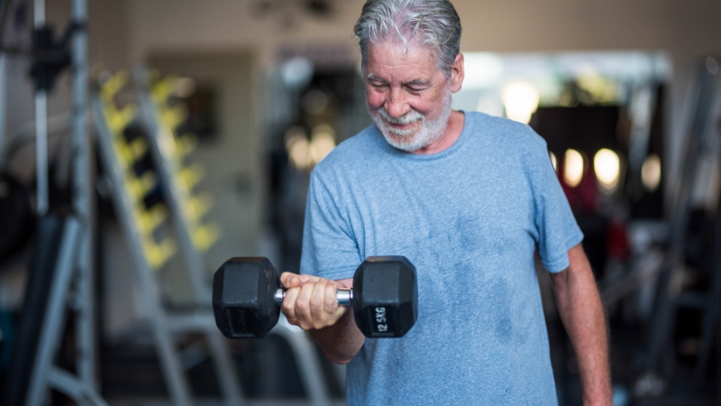 An older man in a blue shirt lifts a dumbbell during a strength training workout at the gym