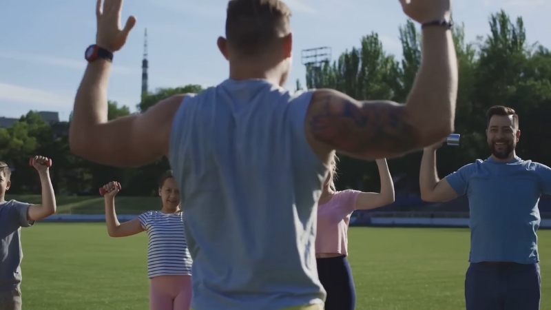A fitness coach leads a group of people exercising together outdoors on a sunny day