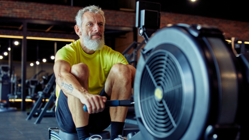 An older man in a bright yellow shirt uses a rowing machine at the gym to maintain strength and endurance