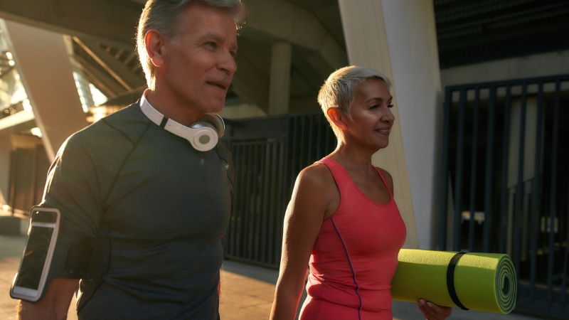An active older couple walks together, with the woman carrying a yoga mat