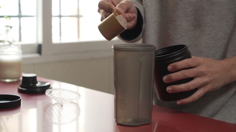 Person scooping cocoa powder into a shaker cup, preparing a nutritional drink