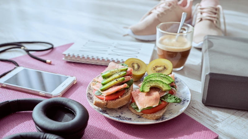 A plate with avocado and ham sandwiches, fresh kiwi, and coffee placed on a yoga mat beside workout gear and sneakers