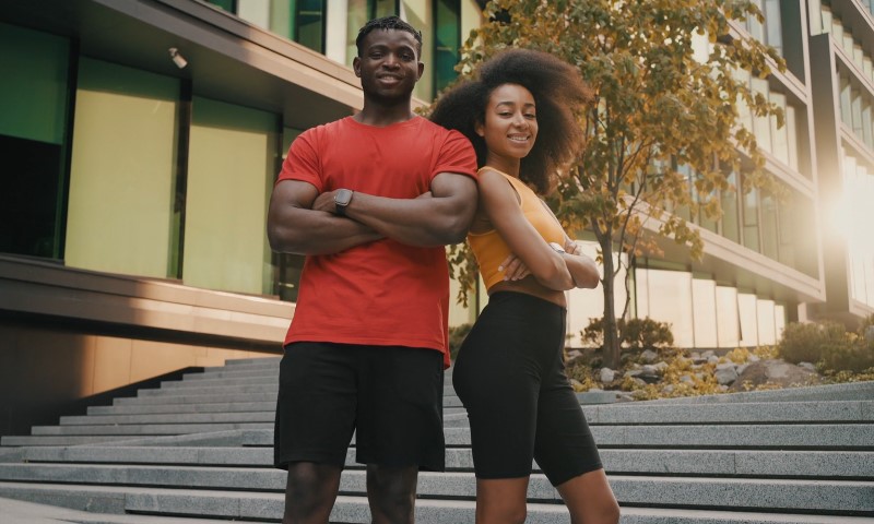 A smiling man in a red shirt and a woman in a yellow top stand back-to-back with arms crossed