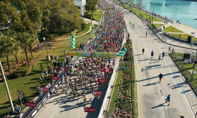 Aerial view of a colorful marathon with hundreds of runners on a sunny street beside a river