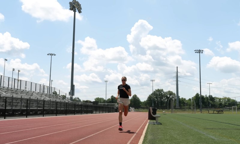 A person runs on an outdoor track under a bright blue sky with fluffy clouds
