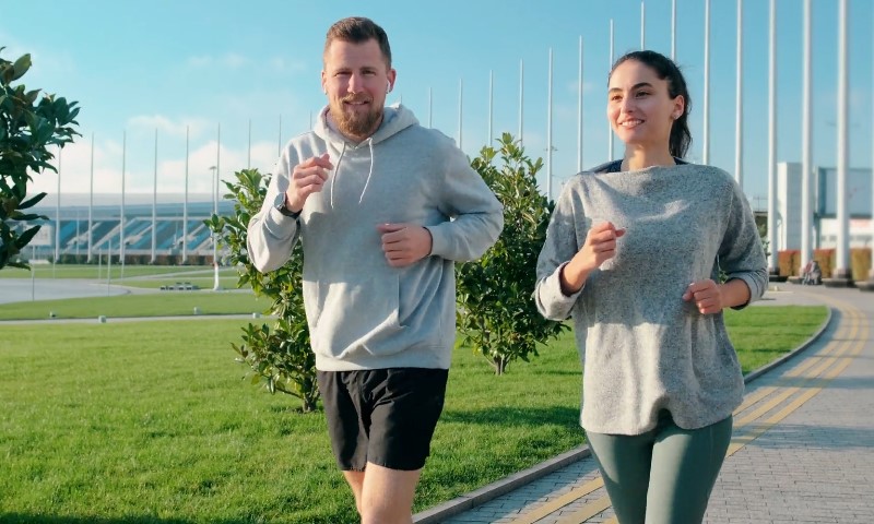 A man and woman jog outside on a sunny day, smiling and wearing casual athletic wear