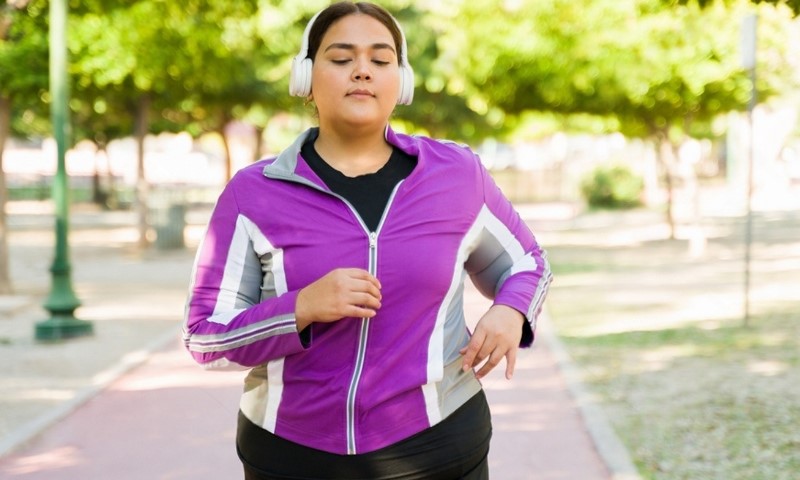 A woman in a purple jacket jogs on a sunny park path, wearing headphones